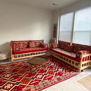 May include: Two red and white patterned couches with wooden frames and red cushions, a brown coffee table, and a red and white patterned rug. The couches are arranged in a living room setting with a white wall and a window with white blinds in the background.
