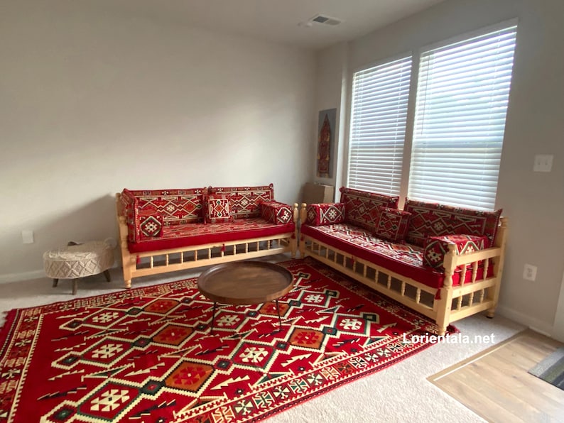 May include: Two red and white patterned couches with wooden frames and red cushions, a brown coffee table, and a red and white patterned rug. The couches are arranged in a living room setting with a white wall and a window with white blinds in the background.