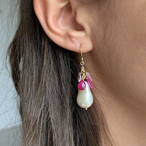 Peut inclure: Une paire de boucles d'oreilles en or avec une grosse perle blanche et des accents de pierres précieuses roses et rouges.
