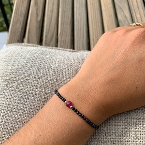 Peut inclure: Un bracelet délicat avec des perles noires et un seul accent de pierre précieuse rose. Le bracelet est porté au poignet.