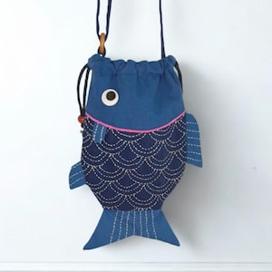 May include: A fish-shaped drawstring bag made of blue fabric with a dark blue lower section featuring white stitched scale patterns. The bag includes a single round eye, a pink trim line, side fins, a tail, and a thin blue shoulder strap.