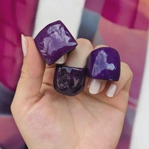 May include: Three dark purple rings with a glossy finish. The rings are different shapes, including a square and a rounded square. The background features a colorful, abstract design with shades of pink, purple, and white.