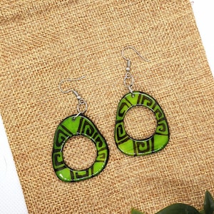 May include: A pair of green and black geometric patterned earrings with silver hooks. The earrings are displayed on a brown burlap bag with green leaves.