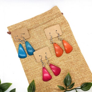 May include: Three pairs of teardrop-shaped earrings in blue, orange, and pink. Each pair is attached to a small, brown card with a silver hook. The earrings are displayed on a burlap bag, with green leaves in the foreground.