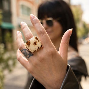 May include: A square ring featuring a smooth, cream-colored surface with irregular dark brown, marbled inclusions. The ring has a metallic band and a flat, geometric face that sits prominently on the finger, showcasing a distinct contrast between the light base and the dark, organic-looking patterns within the material.