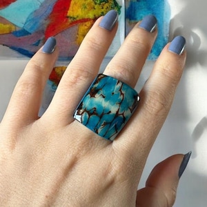 May include: A hand wearing a blue and brown rectangular ring with a marbled pattern. The ring is on the middle finger of the hand.