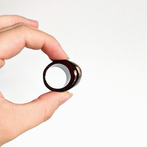 May include: A dark brown wooden ring with a light brown wood grain detail. The ring is held in a hand.
