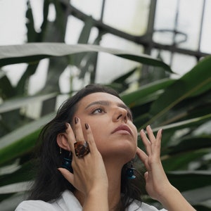 May include: A woman with long dark hair wearing a white shirt and tortoise shell ring. She is looking up and has her hands near her face.