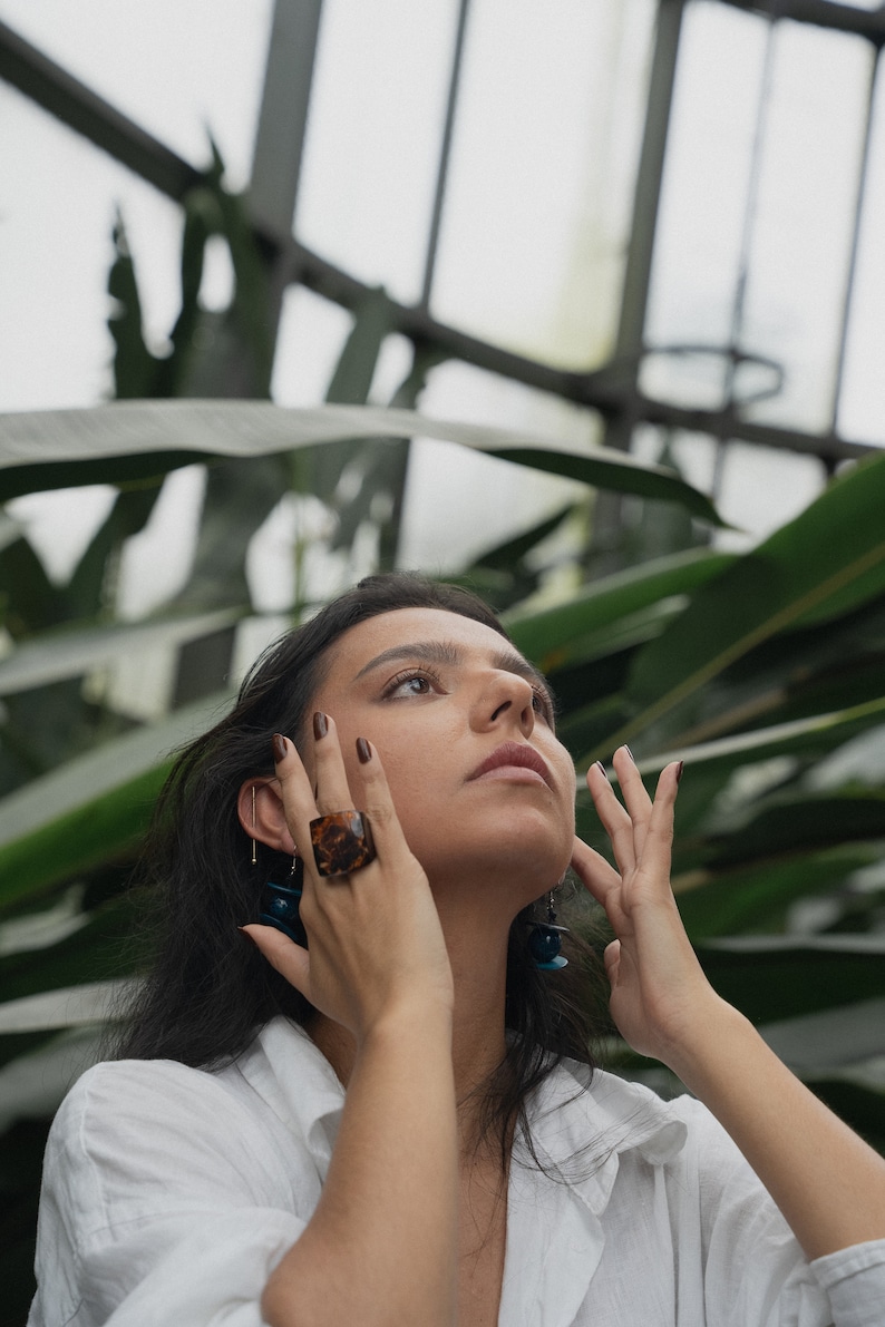 May include: A woman with long dark hair wearing a white shirt and tortoise shell ring. She is looking up and has her hands near her face.