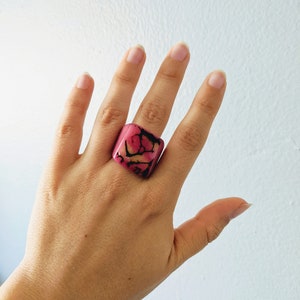 May include: A square pink ring with a marbled design in shades of pink, orange, and black.
