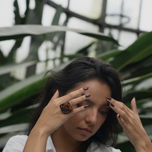 May include: A woman wearing a white shirt with a large, brown, square-shaped ring on her finger. Her nails are painted a dark brown color.
