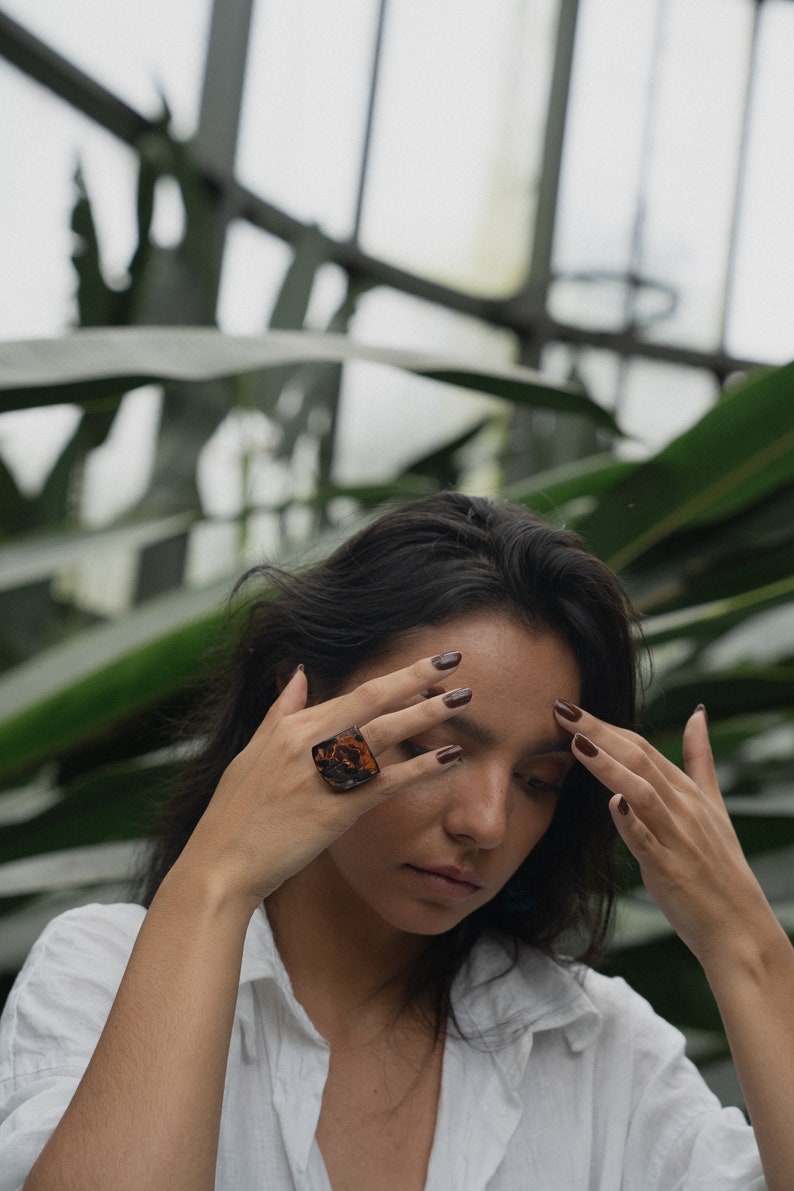 May include: A woman wearing a white shirt with a large, brown, square-shaped ring on her finger. Her nails are painted a dark brown color.