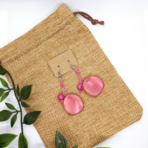 Puede incluir: Pendientes colgantes de color rosa con una cuenta de madera rosa y un dije ovalado rosa. Los pendientes se muestran en una bolsa de arpillera marrón.
