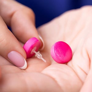 Puede incluir: Un par de pendientes de botón de color rosa brillante. Un pendiente se sostiene, mostrando el poste y el respaldo. El otro pendiente descansa en la palma de una mano. Los pendientes son redondos y brillantes, con un color sólido vibrante.