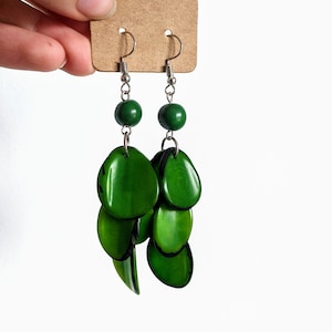 Puede incluir: Un par de pendientes verdes colgantes con una cuenta redonda verde en la parte superior y múltiples piezas verdes en forma de hoja que cuelgan debajo.