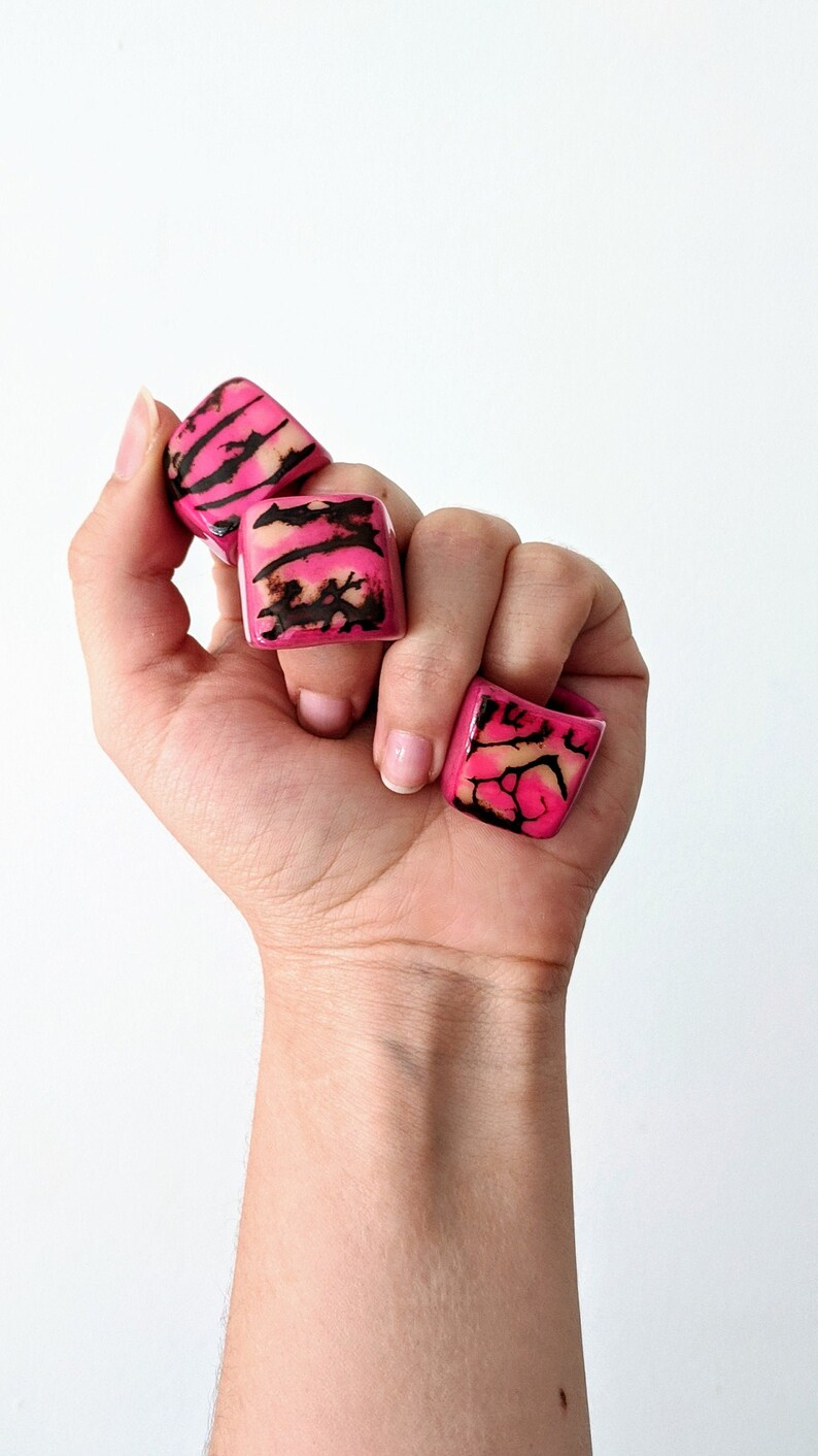 May include: Four pink and black patterned square rings stacked on a hand.