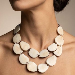 Puede incluir: Un collar de doble hebra con discos circulares de forma irregular, lisos y de color blanco roto. Los discos están conectados por finos eslabones de cordón marrón oscuro, creando un aspecto orgánico y en capas alrededor del cuello.