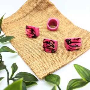 May include: Four pink and black patterned rings on a brown burlap bag. The rings are made of wood and have a unique design.
