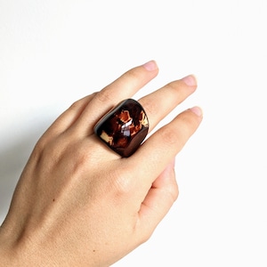 May include: A large, brown and cream resin ring with a unique marbled pattern. The ring is worn on a person's finger.
