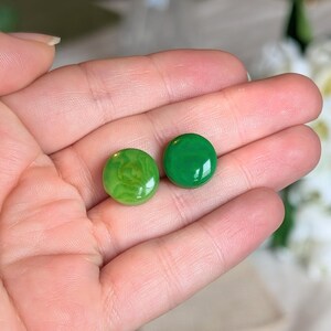 Puede incluir: Dos piedras preciosas redondas y pulidas de color verde. Una es de un verde vibrante y sólido, mientras que la otra tiene un aspecto más claro y veteado. Las piedras preciosas se muestran en una mano, mostrando su color y textura.