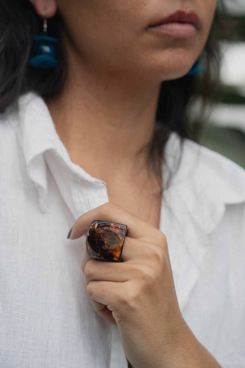 May include: A close-up of a person's hand wearing a large, brown and black resin ring. The ring has a unique marbled pattern.