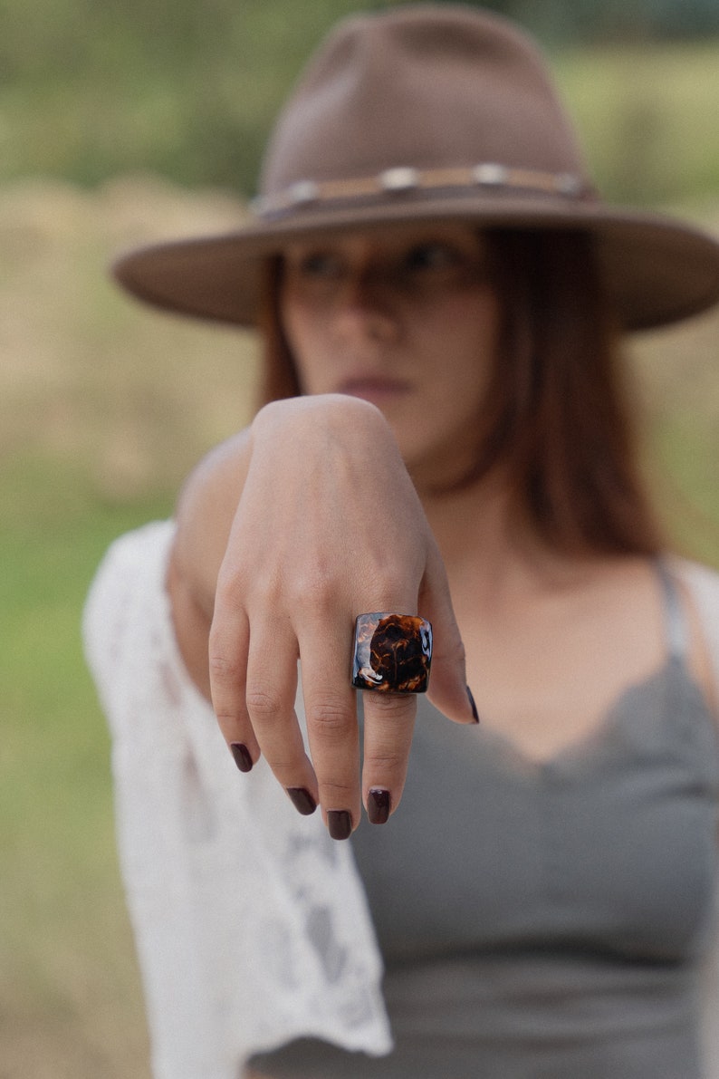 May include: A close-up of a hand wearing a square-shaped ring with a brown and gold marbled design.