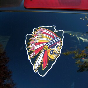 Op de afbeelding: Een kleurrijke sticker met een schedel met een Indiaanse hoofdtooi. De hoofdtooi heeft rode, gele en witte veren, en een blauw-witte kralenband. De schedel is geel en zwart, met een witte omtrek.