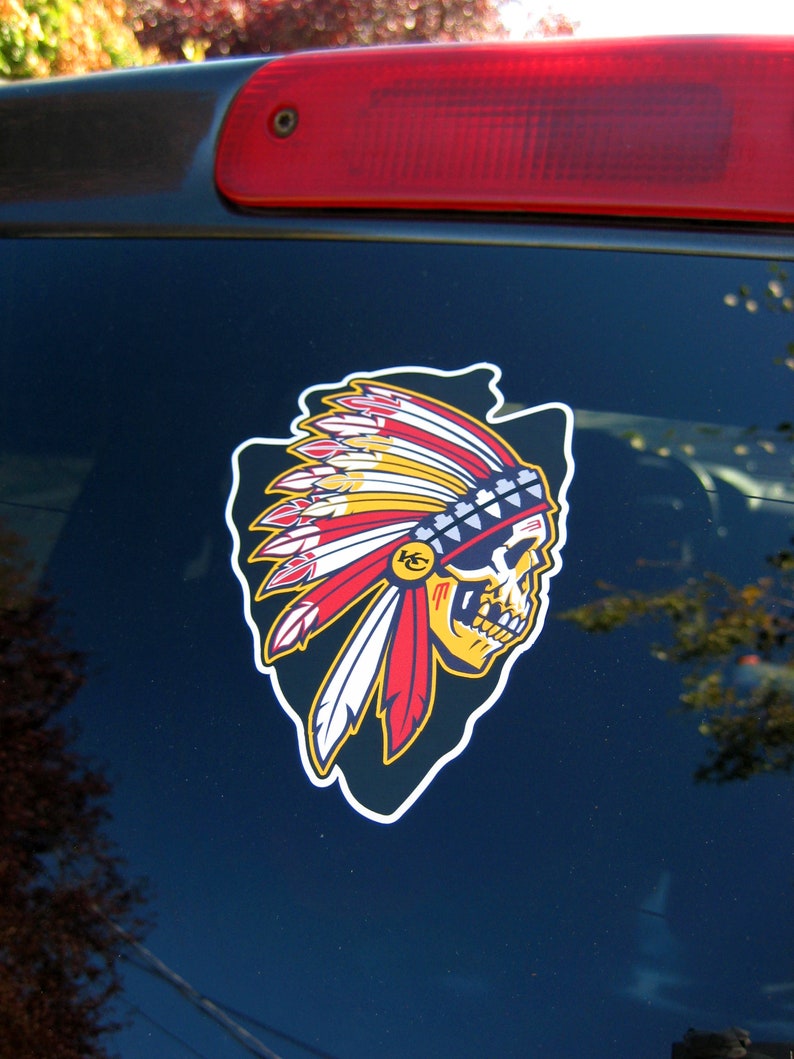 Op de afbeelding: Een kleurrijke sticker met een schedel met een Indiaanse hoofdtooi. De hoofdtooi heeft rode, gele en witte veren, en een blauw-witte kralenband. De schedel is geel en zwart, met een witte omtrek.