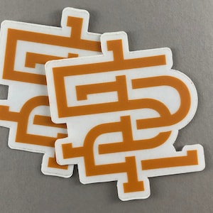 May include: Two white stickers with a repeating geometric design in a warm, golden-brown color. The stickers have a white border and are slightly overlapping, creating a layered effect. The background is a solid, neutral gray.