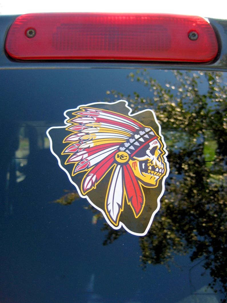 Op de afbeelding: Een sticker met een gestileerde schedel met een Indiaanse hoofdtooi in rood, geel en wit. De sticker heeft de vorm van een pijl en is bevestigd op een donkere ondergrond, met daarboven een rood remlicht.