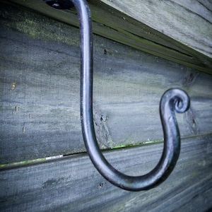 May include: A handcrafted, dark metal hook mounted on weathered wooden planks. The hook has a curved design with a decorative scroll at the end. The wood has a gray-blue hue, and the hook is likely for hanging items.