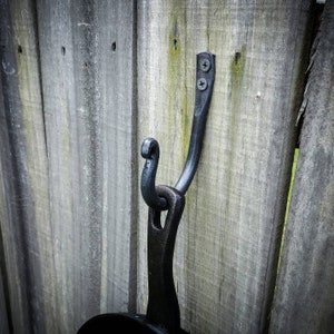 May include: Black metal hook mounted on a wooden fence. The hook is shaped like a bird's head with a curved neck and a pointed beak.