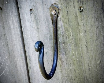 May include: A black metal hook with a rounded top and a single screw. The hook is mounted on a weathered wooden surface.