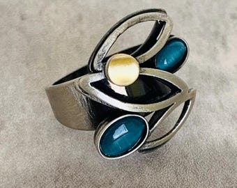 May include: A silver-toned ring with a unique design. It features a gold-colored circular accent and three oval-shaped teal-colored stones. The ring has an open, artistic design with curved lines and a textured band.