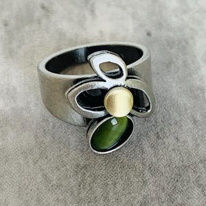 Może przedstawiać: Srebrny pierścionek z wzorem zielonego i złotego kwiatu. Kwiat ma trzy płatki i centralny kamień.