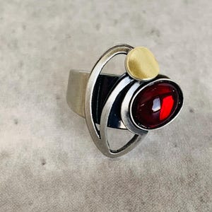May include: A silver ring with a red gemstone and a gold accent. The ring has a modern, abstract design.