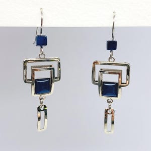 Peut inclure: Paire de boucles d'oreilles argentées au design géométrique. Chaque boucle d'oreille présente un carré bleu foncé en haut, un cadre carré argenté et un carré bleu foncé au centre. Un rectangle ouvert argenté est suspendu en dessous.