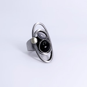 May include: A silver ring with an oval design, featuring two dark gemstones and a small, round silver accent. The ring has a modern, geometric aesthetic, with a polished finish.