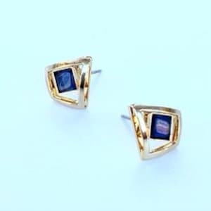 May include: A pair of gold-colored stud earrings with a geometric design. Each earring features a square, dark blue center stone set within a triangular frame. The earrings are displayed on a light blue surface.