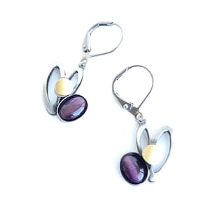 May include: A pair of silver earrings with a distinctive design. Each earring has a purple oval stone, a gold-coloured circle, and a silver heart-shaped element. The earrings have lever-back closures.