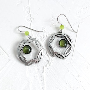May include: Silver earrings with a green stone set in a circular frame. The earrings have a delicate, openwork design and are accented with small green beads.