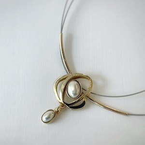 May include: A gold-toned necklace with a central oval pendant and a smaller, teardrop-shaped pendant. The design incorporates black enamel accents and two strands of gray wire. The necklace features a large, white, oval-shaped pearl.