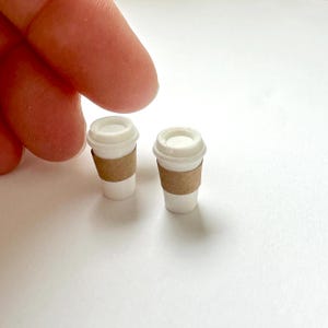 May include: Two miniature white coffee cups with brown cardboard sleeves. The cups have white lids and are set against a white background. The cups are small, likely for a dollhouse or miniature collection.