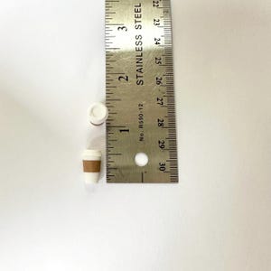May include: Miniature white coffee cup with a brown sleeve and a white lid. The cup is approximately 2 cm tall. A stainless steel ruler is next to the cup for scale.