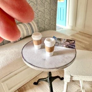 May include: A miniature scene with two small white coffee cups with brown sleeves on a round table. The table has a marble effect top and a black, ornate base. A magazine rests on the table. The scene is set in a dollhouse.