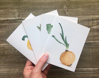 May include: A hand holds a stack of four white greeting cards. Each card features a watercolour illustration of vegetables: a yellow squash, a sprig of herbs, a green leafy vegetable, and a brown onion with green sprouts.