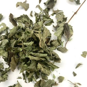 May include: Close-up of dried green leaves and stems scattered on a white surface. The leaves have a textured appearance, with visible veins and a muted green color. Some leaves are whole, while others are fragmented.