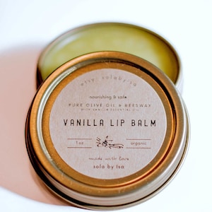 May include: A gold metal tin with a brown label that reads "etsy solaby'sa nourishing & safe PURE OLIVE OIL & BEESWAX WITH VANILLA ESSENTIAL OIL VANILLA LIP BALM 1 oz organic made with love sola by Isa".