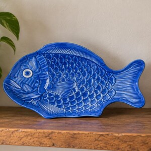May include: A vibrant cobalt blue ceramic serving platter shaped like a fish. The platter features detailed embossed scales, fins, and a prominent eye. The surface has a glossy finish that reflects light, highlighting the intricate texture of the fish design.