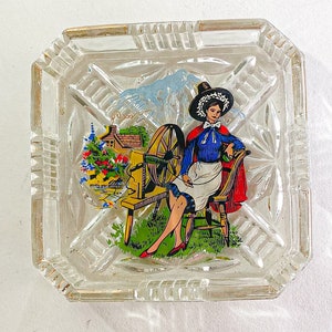 May include: A clear glass ashtray with a raised design around the edge. The ashtray features a colourful image of a woman in a red and blue dress sitting on a chair with a spinning wheel. The scene is set in a rural setting with a small house and trees.